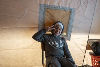 A smiling young child in a gray tracksuit sits on a chair inside a fabric tent, making a peace sign with her hand. Soft light filters through the tent walls behind her.