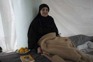 An older woman dressed in black sits beside a bed inside a tent-like shelter, holding a folded blanket. A small plate of fruit and a book rest on the bed next to her.