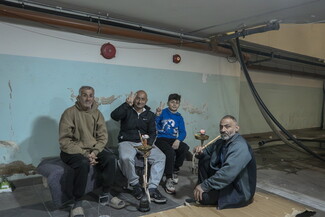Three adult men and one teenage boy sitting together in a modest shelter-like room with exposed pipes and peeling paint, sharing hookah (shisha). Two make victory signs toward the camera. They wear hoodies and jackets, relaxed yet resilient mood, low-lit environment with visible water stains and worn walls, social gathering in confined space.