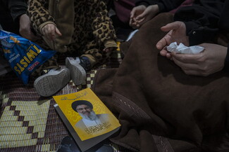 A close-up view of people sitting on a woven mat, including a child in a patterned jacket. One person holds tissues in their hands, while a yellow-covered book with Arabic text lies on the floor nearby next to a bag of snacks. Their palms are turned upward in prayer.
