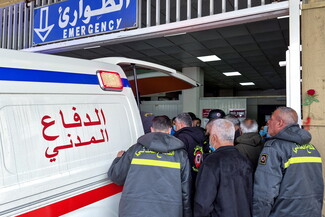 Emergency hospital entrance with Arabic and English “Emergency” signage, paramedics and civil defense personnel gathered at the open rear of an ambulance, first responders in uniforms and face masks assisting with patient transfer, urgent healthcare setting, hospital exterior.