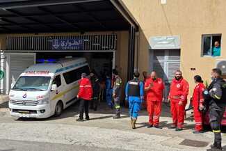 Ambulance parked outside hospital emergency department with Arabic “Emergency” sign and “Corona Section” notice visible, multiple paramedics in red and blue uniforms standing and walking near the entrance, healthcare workers coordinating response, bright daylight, emergency medical services scene.