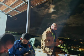 Three men sit and stand on a covered patio at night; two use their phones while the third stands with a cigarette, with a cloudy moonlit sky in the background.