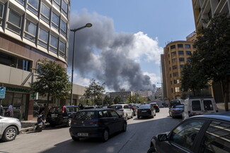 Thick, dark smoke rises into the sky from buildings in the distance, viewed from a city street with cars parked and driving along both sides.