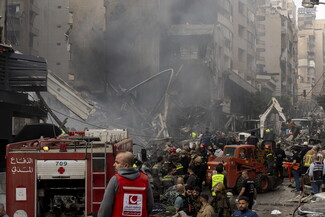 Emergency responders and firefighters work amid rubble and smoke in a city street following a building collapse or explosion, with damaged vehicles and debris visible.