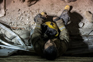 A tired firefighter sits on rubble, holding a yellow helmet, with dirty boots and gloves.