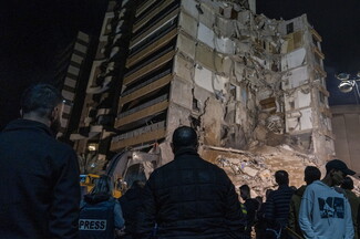 Crowd gathers at night before a partially collapsed building, with exposed interiors and debris. Somber scene with press and onlookers observing.