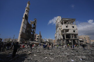 A  photograph of a destroyed mosque. People and rescue workers and first responders stand amid the rubble, dwarfed by the size of the building and its destruction. In the background, the sky is cloudy but blue.