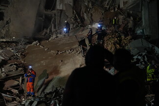 A nighttime photograph of a destroyed building. Rescue workers and first responders stand amid the rubble, dwarfed by the size of the building and its destruction.