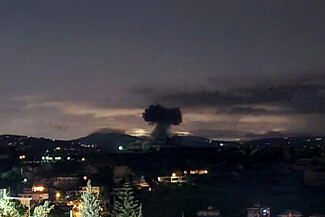 A dark plume of smoke rises into the night sky above a distant city, with lights from buildings visible in the foreground.