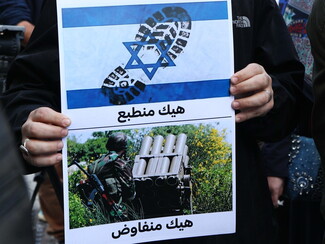 A demonstrator carries a sign that reads: “This is how we normalize” and “This is how we negotiate.” The former is written under an Israeli flag with a boot print stepping on the flag. The latter is written underneath a photo of someone standing next to a rocket/missile launcher.