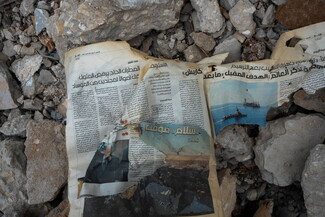 Torn newspaper in Arabic with a photo of a ship, lying on a pile of rubble and stones.