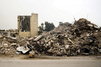 A large pile of rubble from destroyed buildings fills the foreground. In the background, a mural of a uniformed man is painted on a tall, standing wall.