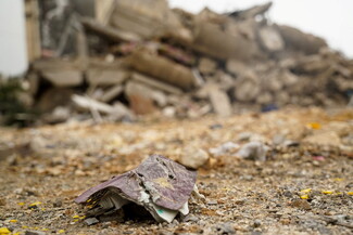 Tattered papers lie on rocky ground with a collapsed building in the background,