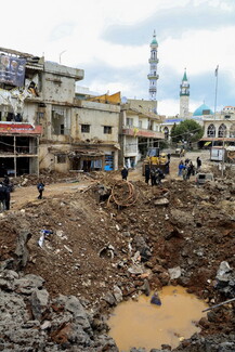 Large crater filled with muddy water in a street surrounded by damaged buildings and debris, with people inspecting the area; mosque minarets visible in the background.