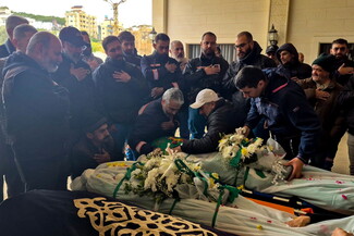 A solemn group of people gathers around a covered body, adorned with white flowers. 