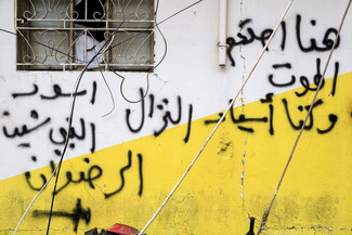 Graffiti in Arabic covers a white and yellow exterior wall beneath a barred window with exposed wires hanging across the scene.
