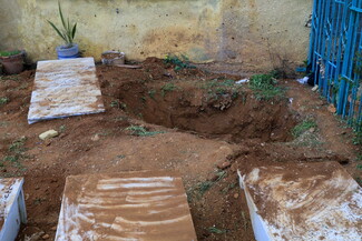 A freshly dug grave sits in a bare, dirt-covered area, surrounded by simple headstones. 