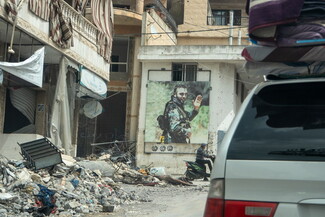 Street scene with rubble and debris in front of damaged buildings after Israeli strikes. A large portrait of a man in military attire hangs on a wall.