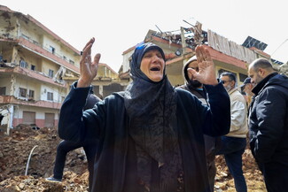 A distressed woman in a hijab stands with arms raised amidst rubble and damaged buildings.