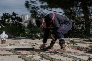 A paramedic in a dark uniform with red stripes kneels and spray-paints on a concrete surface of a grave at a cemetery.