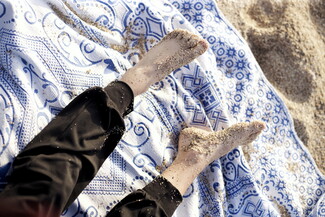 A child's bare feet covered in sand resting on a blue and white patterned blanket at the beach. The scene conveys relaxation and a casual, sunny atmosphere.