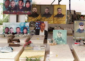 A cemetery scene featuring gravestones adorned with flowers, framed photos of martyrs, and decorative tributes. Portraits of various individuals, both fighters and civilians, are displayed above the graves, highlighting a somber and respectful atmosphere.
