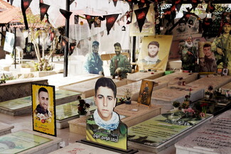 A cemetery scene featuring gravestones adorned with flowers, framed photos of martyrs, and decorative tributes. Portraits of various individuals, both fighters and civilians, are displayed above the graves, highlighting a somber and respectful atmosphere.