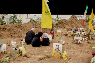 Three people sit on the ground, facing gravestones adorned with flowers and portraits. Yellow flags flutter above. The mood is somber.