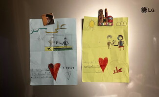Two children's drawings on a metallic surface. The left is drawn on blue paper, and shows a flag and hearts with text that reads: “Ali Samra. I am Zein. I love you baba, and you are my kind and loving father.” It shows a child giving flowers to his father. The drawing on the right is drawn on yellow paper, has a sun and hearts, and shows a child giving flowers to his mother. It reads:  “Ola. I love you mama, and you are my kind and sweet mother.”