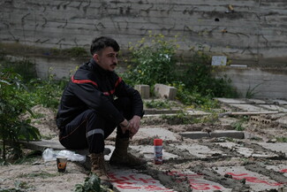 A paramedic sits on a dirt path, looking pensive. Around him are plants, a spray can and a coffee cup as he overlooks the grave of his friend in a mass cemetery. 