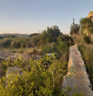 A weathered stone wall with a house appearing in the distance under a bright sky.