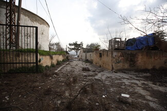 A narrow, muddy path between weathered concrete walls is flanked by a rusty gate and graffiti.
