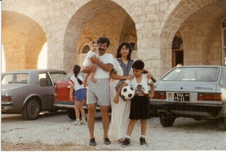 A family of four stands smiling in front of a stone building with arches. The father holds a baby, the mother stands beside them, and a boy holds a soccer ball.