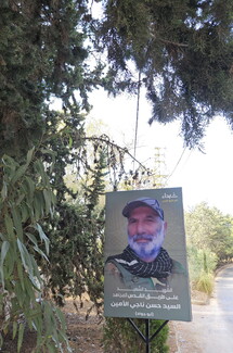 A memorial sign featuring a martyred man with a beard and cap, set against a backdrop of trees and a dirt path.
