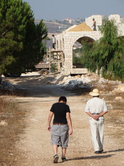 Two people walk on a dirt path towards an under-construction stone building in a rural landscape. 
