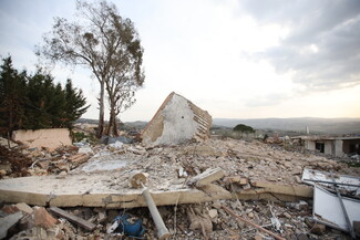 Rubble and debris scattered across a barren landscape, with a partially collapsed building and a lone tree under a cloudy sky.
