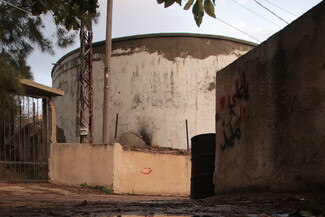 A weathered, cylindrical large water tank with peeling paint stands behind a rough concrete wall. 