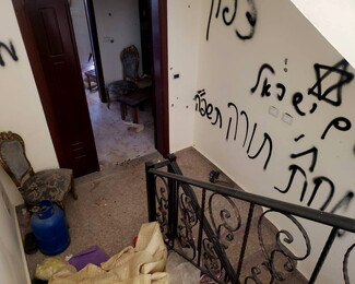 An interior view looking down a staircase inside a damaged and messy home. The white wall next to the stairs is covered in large Hebrew graffiti spray-painted in black, including a Star of David. The landing and adjacent rooms are in disarray, littered with a blue gas cylinder, bundled blankets, loose trash, and displaced classic-style wooden chairs.