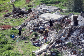 A woman navigates through a massive pile of concrete rubble on a grassy hillside while a man watches nearby. An overturned white vehicle is visible in the wreckage.