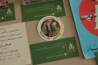 A close-up of archival display items, featuring a circular white sticker showing a soldier in camouflage facing an elderly man, surrounded by green descriptive labels containing Arabic text.