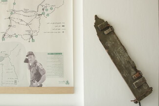 A wall display showing the edge of a historical map with a portrait of a military official on the left, next to a piece of green military metal debris with Hebrew stencil lettering mounted on the right.