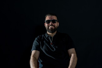 A photo of a smiling man in front of a black background. He is bearded, wearing black, and wearing sunglasses. There are scars on his face.
