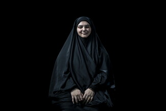 امرأة مبتسمة ترتدي حجاباً أسود وتجلس أمام خلفية سوداء. على وجهها ويديها ندوب واضحة.