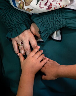 A close-up, top-down photograph focusing on the hands of an adult and a young child resting against a dark green fabric background. The adult's hand, wearing a gold ring with a large oval red gemstone surrounded by small diamonds, shows healed amputations at the fingertips. A child's smaller hands are gently placed over the adult's hand in a comforting, tender gesture. The adult is wearing a dark green garment with tightly smocked, ruffled cuffs.