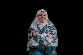 A medium studio portrait of a woman, Rawan al-Moussawi, seated against a solid black background. She is wearing a white hijab patterned with vibrant blue, purple, and orange flowers, along with a dark green long-sleeved dress. She is smiling warmly with her eyes closed. Visible scarring and healing shrapnel injuries are present across her face, particularly on her forehead, nose, and cheeks.