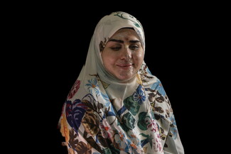 A medium studio portrait of a woman, Rawan el-Moussawi, seated against a solid black background. She is wearing a white hijab patterned with vibrant blue, purple, and orange flowers, along with a dark green long-sleeved dress. She is smiling warmly with her eyes closed. Visible scarring and healing shrapnel injuries are present across her face, particularly on her forehead, nose, and cheeks.