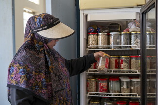 A woman wearing a floral hijab and a wide-brimmed sun visor pulls a glass jar of seeds from a refrigerator neatly stocked with numerous jars of preserved seeds, beans, and grains.