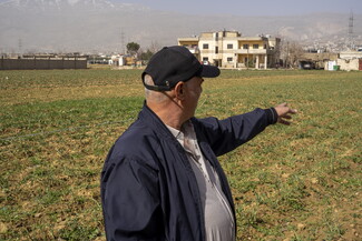 A farmer wearing a black cap and navy jacket stands in a field of young green crops, pointing his right arm toward distant buildings and hazy mountains.