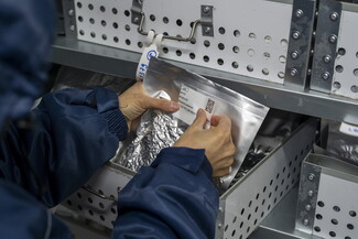 A close-up shot of a person in a heavy winter coat retrieving a labeled, silver metallic foil seed pouch from a white perforated storage drawer.